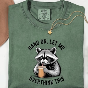 May include: Sage green t-shirt featuring a raccoon graphic with a drink and the text "Hang on, let me overthink this." A gold star pendant hangs from a gold chain. The shirt is a Comfort Colors brand.