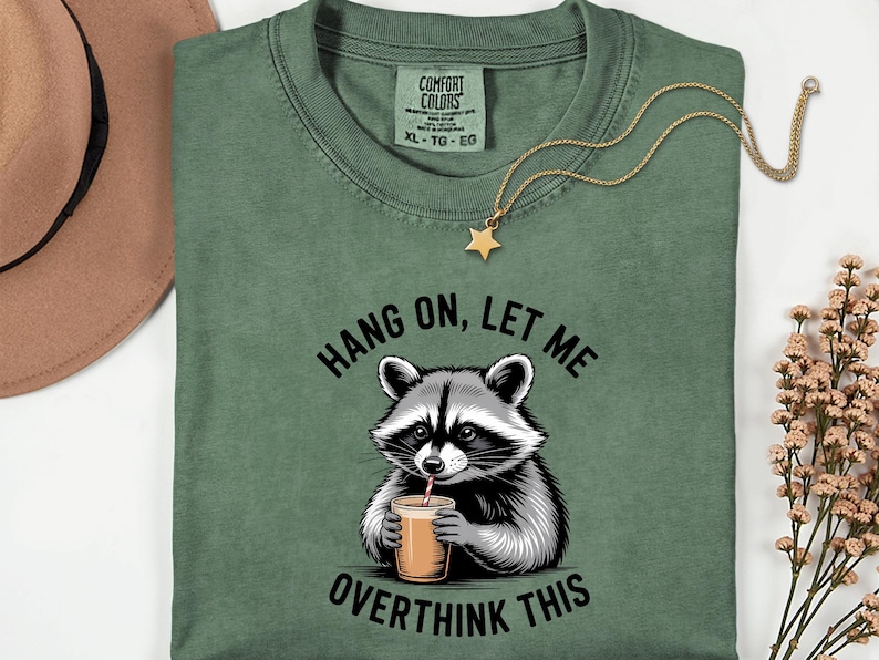 May include: Sage green t-shirt featuring a raccoon graphic with a drink and the text "Hang on, let me overthink this." A gold star pendant hangs from a gold chain. The shirt is a Comfort Colors brand.