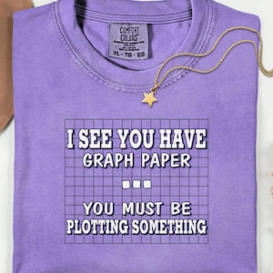 May include: A lavender Comfort Colors t-shirt with the text "I SEE YOU HAVE GRAPH PAPER YOU MUST BE PLOTTING SOMETHING" in white. A gold star pendant necklace rests on the shirt. A brown hat is visible in the background.