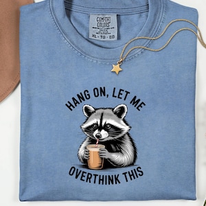 May include: A pale blue t-shirt featuring a raccoon illustration and the phrase "Hang on, let me overthink this". A gold star pendant necklace is draped over the shirt. The shirt has a Comfort Colors label.