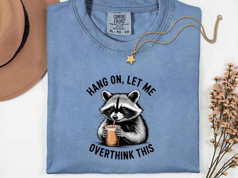 May include: A pale blue t-shirt featuring a raccoon illustration and the phrase "Hang on, let me overthink this". A gold star pendant necklace is draped over the shirt. The shirt has a Comfort Colors label.