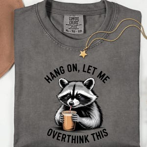 May include: Dark grey t-shirt featuring a raccoon illustration drinking from a cup, with the text "HANG ON, LET ME OVERTHINK THIS". A gold star pendant necklace and a brown hat are also visible.