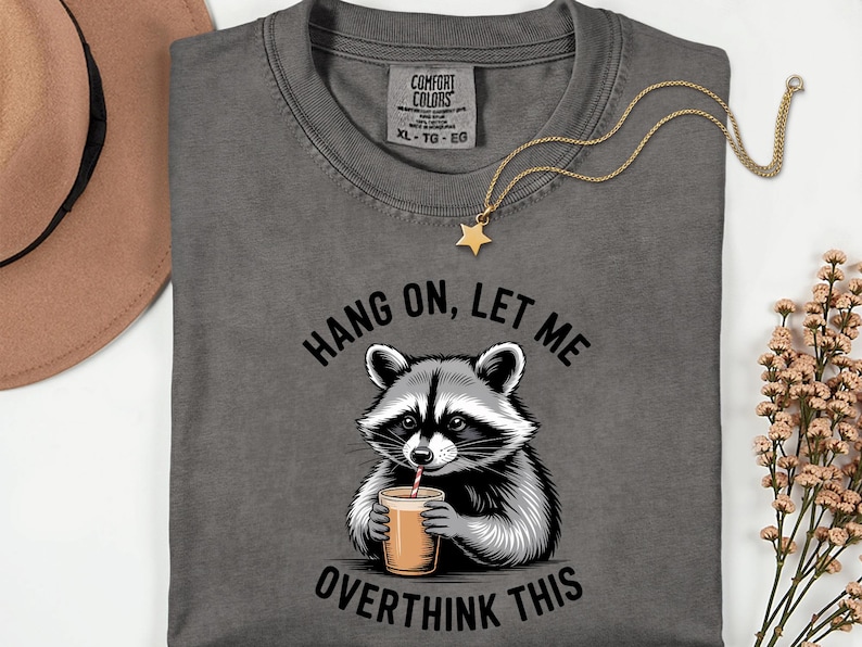 May include: Dark grey t-shirt featuring a raccoon illustration drinking from a cup, with the text "HANG ON, LET ME OVERTHINK THIS". A gold star pendant necklace and a brown hat are also visible.