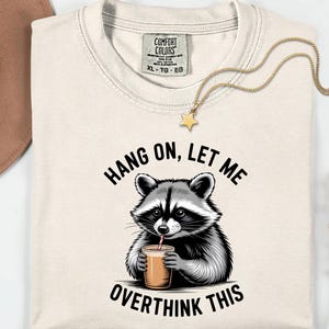 May include: A cream-coloured t-shirt with a raccoon illustration drinking from a cup with a straw. The text "HANG ON, LET ME OVERTHINK THIS" is above and below the raccoon. A gold necklace with a star pendant and a brown hat are also visible.