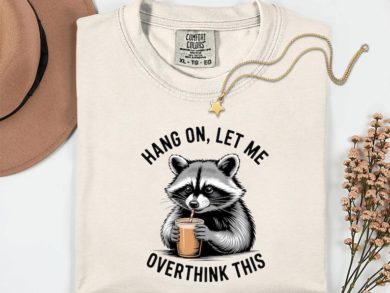 May include: A cream-coloured t-shirt with a raccoon illustration drinking from a cup with a straw. The text "HANG ON, LET ME OVERTHINK THIS" is above and below the raccoon. A gold necklace with a star pendant and a brown hat are also visible.