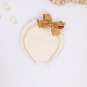May include: A beige headband with a gold-colored band and a tan bow. The headband is on a round, cream-colored base. The image is on a white background with decorative wooden beads and small, light green pom-poms.