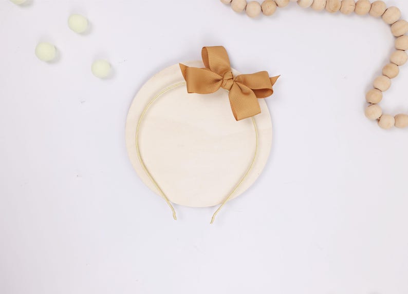 May include: A beige headband with a gold-colored band and a tan bow. The headband is on a round, cream-colored base. The image is on a white background with decorative wooden beads and small, light green pom-poms.