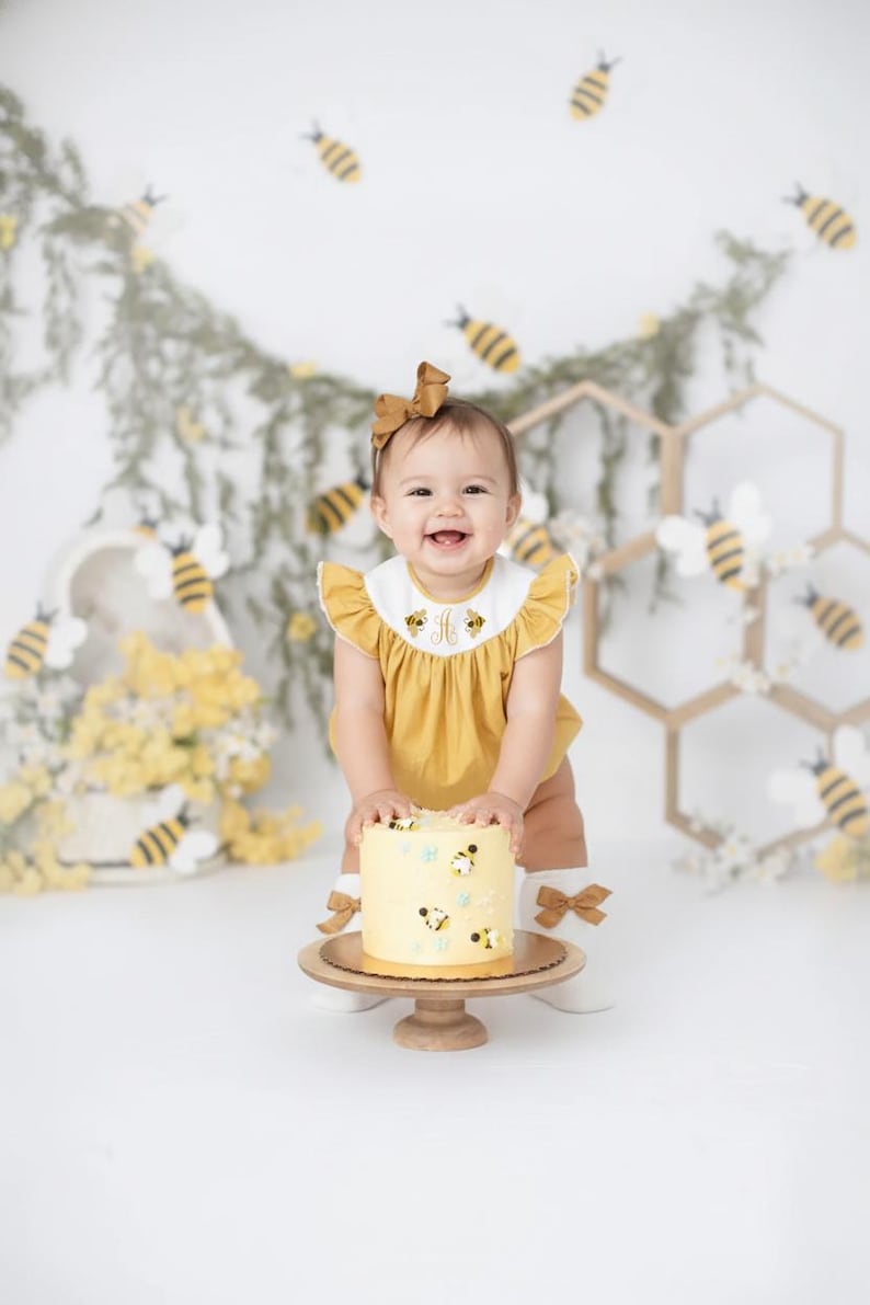 May include: A smiling baby in a yellow outfit with a matching bow, holding a cake decorated with bees. The cake sits on a wooden stand. The background includes bee decorations and honeycomb-shaped structures.