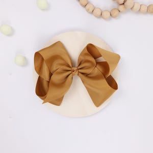 May include: A large, golden-brown hair bow made of grosgrain ribbon. The bow is centered on a light-colored, round wooden base. Decorative elements include small, pale green pom-poms and a string of wooden beads.