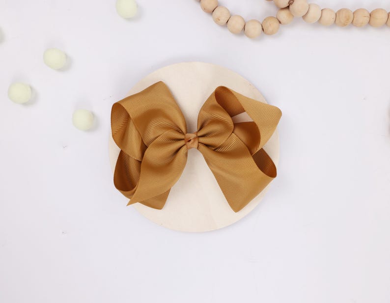 May include: A large, golden-brown hair bow made of grosgrain ribbon. The bow is centered on a light-colored, round wooden base. Decorative elements include small, pale green pom-poms and a string of wooden beads.
