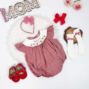 May include: A red and white gingham baby romper with matching headband, socks, and shoes. The romper has embroidered strawberries and the name "Jessica". A wooden "MOM" sign and red bows complete the ensemble.