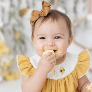May include: A baby wearing a mustard yellow dress with a white bib featuring a bee and the letter "A". The baby has a matching bow in their hair and is eating cake, with frosting on their face.