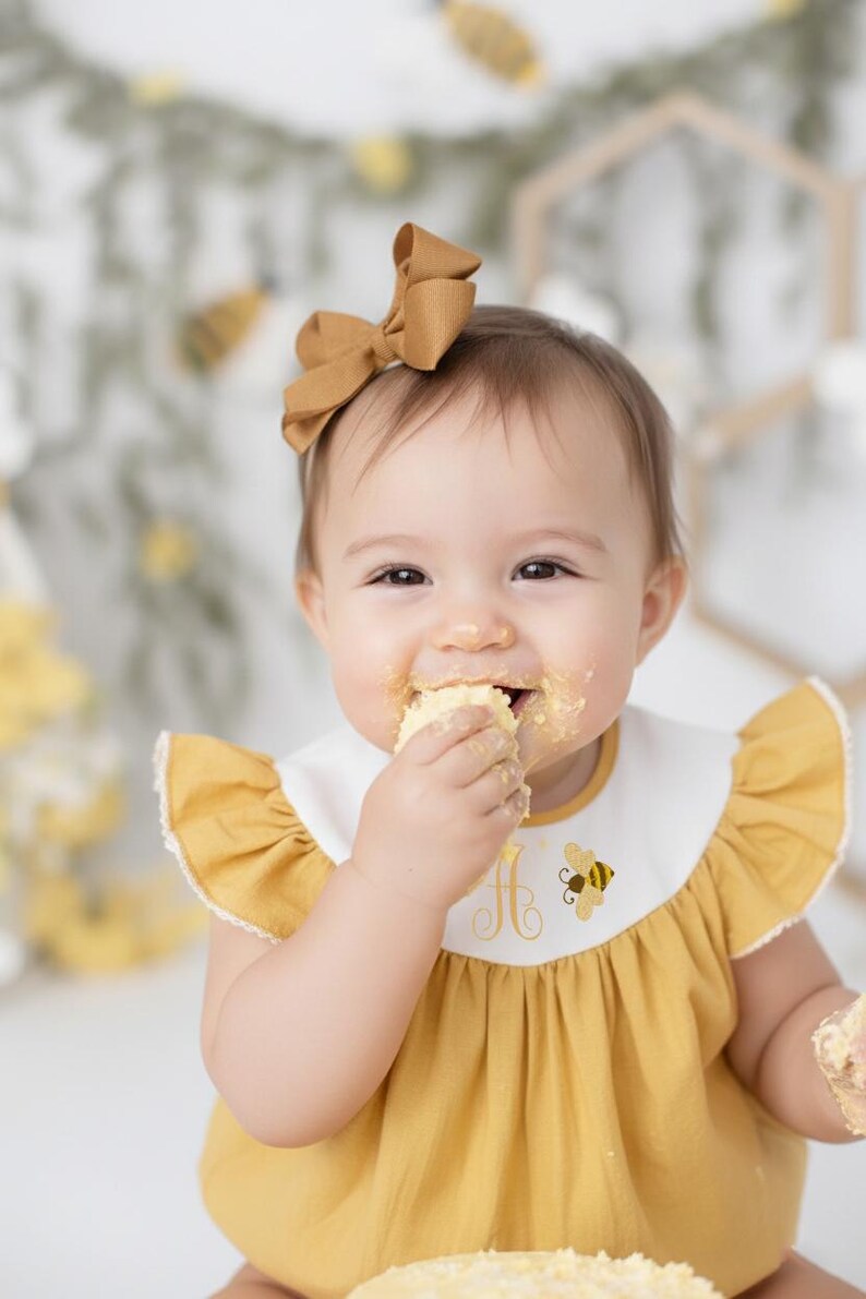 May include: A baby wearing a mustard yellow dress with a white bib featuring a bee and the letter "A". The baby has a matching bow in their hair and is eating cake, with frosting on their face.