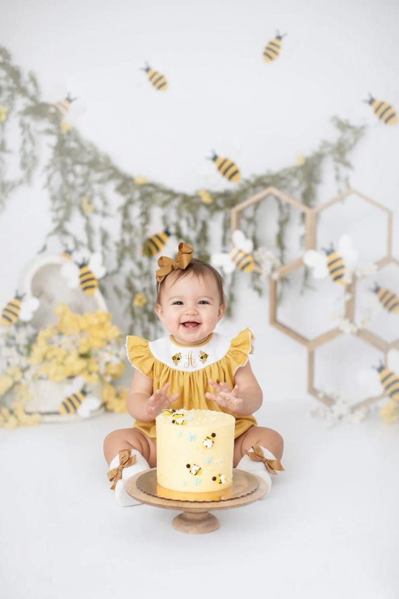 May include: A smiling baby wearing a yellow romper with a white collar, decorated with bees, sits in front of a cake. The cake is yellow and decorated with bees. The background includes bee decorations and a honeycomb.