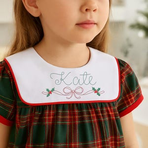 Puede incluir: Vestido a cuadros verdes, rojos y blancos con cuello blanco. El cuello está ribeteado en rojo y bordado con el nombre "Kate" en cursiva, junto con un lazo y ramitas de acebo. El vestido tiene mangas cortas.