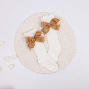 May include: A pair of white knee-high socks with a large, light brown bow at the top of each sock. The socks are displayed on a cream-colored, woven mat against a white background. The bows add a decorative touch.