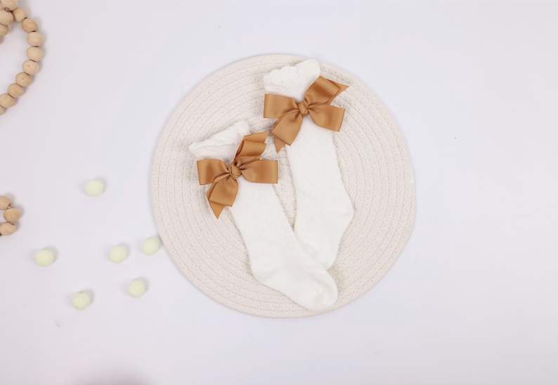 May include: A pair of white knee-high socks with a large, light brown bow at the top of each sock. The socks are displayed on a cream-colored, woven mat against a white background. The bows add a decorative touch.