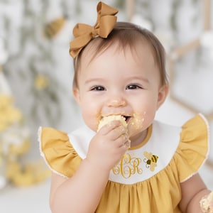 May include: A baby wearing a mustard yellow dress with a white bib featuring a gold embroidered monogram and a bee graphic. The baby has a matching bow in their hair and is eating a piece of cake.