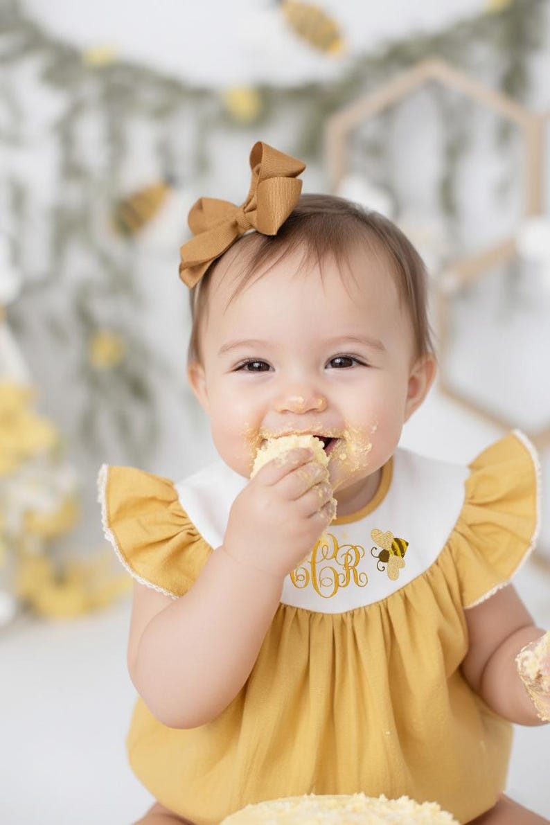 May include: A baby wearing a mustard yellow dress with a white bib featuring a gold embroidered monogram and a bee graphic. The baby has a matching bow in their hair and is eating a piece of cake.