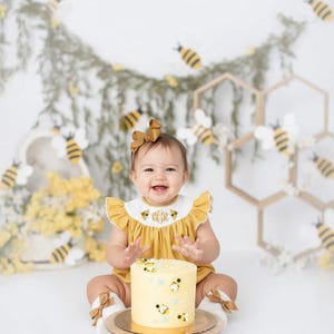 May include: A smiling baby in a yellow romper with a matching bow, sitting in front of a cake decorated with bees. The cake is on a wooden stand. The background includes bee decorations and floral accents, creating a cheerful, celebratory scene.