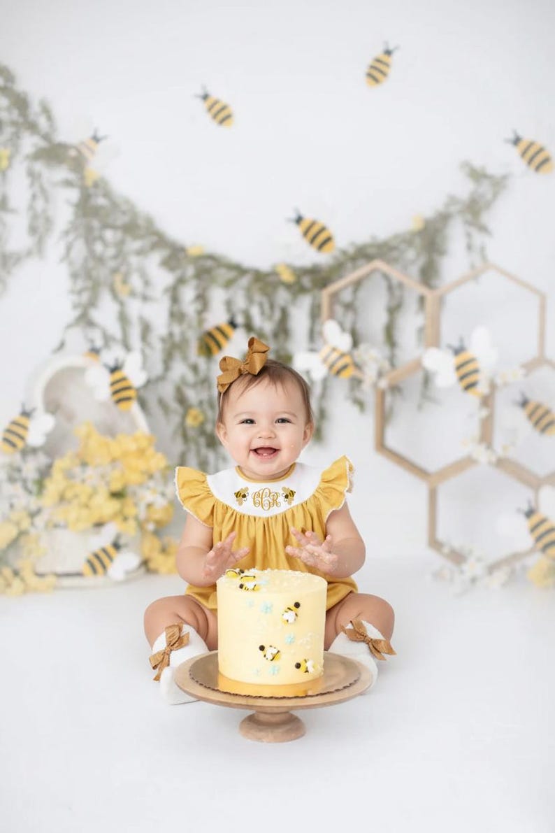May include: A smiling baby in a yellow romper with a matching bow, sitting in front of a cake decorated with bees. The cake is on a wooden stand. The background includes bee decorations and floral accents, creating a cheerful, celebratory scene.