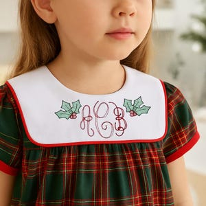 May include: A green, red, and white plaid dress with a white collar. The collar is trimmed in red and features embroidered holly leaves and a monogram. The dress has short sleeves with red trim.