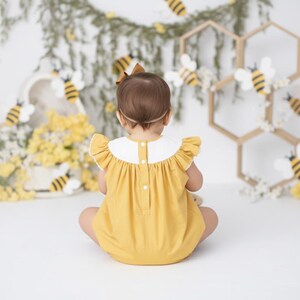 May include: A yellow romper with a white collar and ruffle sleeves. The background features bee decorations, honeycomb-shaped structures, and floral arrangements, creating a whimsical, spring-themed setting.