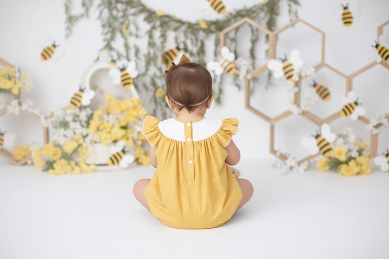 May include: A yellow romper with a white collar and ruffle sleeves. The background features bee decorations, honeycomb-shaped structures, and floral arrangements, creating a whimsical, spring-themed setting.