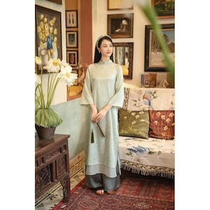 Op de afbeelding: Een lichtgroene jurk met lange mouwen, een mandarijnkraag en een gelaagde rok. De jurk wordt gedragen met een bijpassende broek met wijde pijpen. De vrouw houdt een decoratieve waaier vast. De achtergrond omvat ingelijste kunstwerken en een bank met bloemenpatroon.