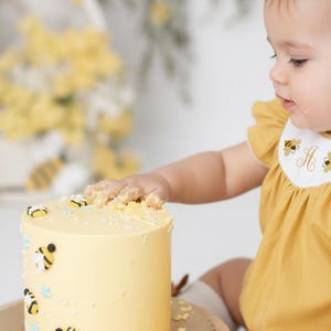 May include: A yellow cake decorated with bee-themed fondant decorations. The cake sits on a wooden cake stand. A child in a yellow outfit reaches for the cake. The cake is decorated with small bees and blue star-shaped sprinkles.