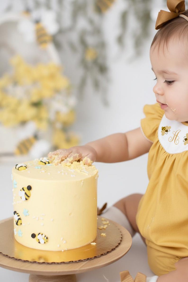 May include: A yellow cake decorated with bee-themed fondant decorations. The cake sits on a wooden cake stand. A child in a yellow outfit reaches for the cake. The cake is decorated with small bees and blue star-shaped sprinkles.