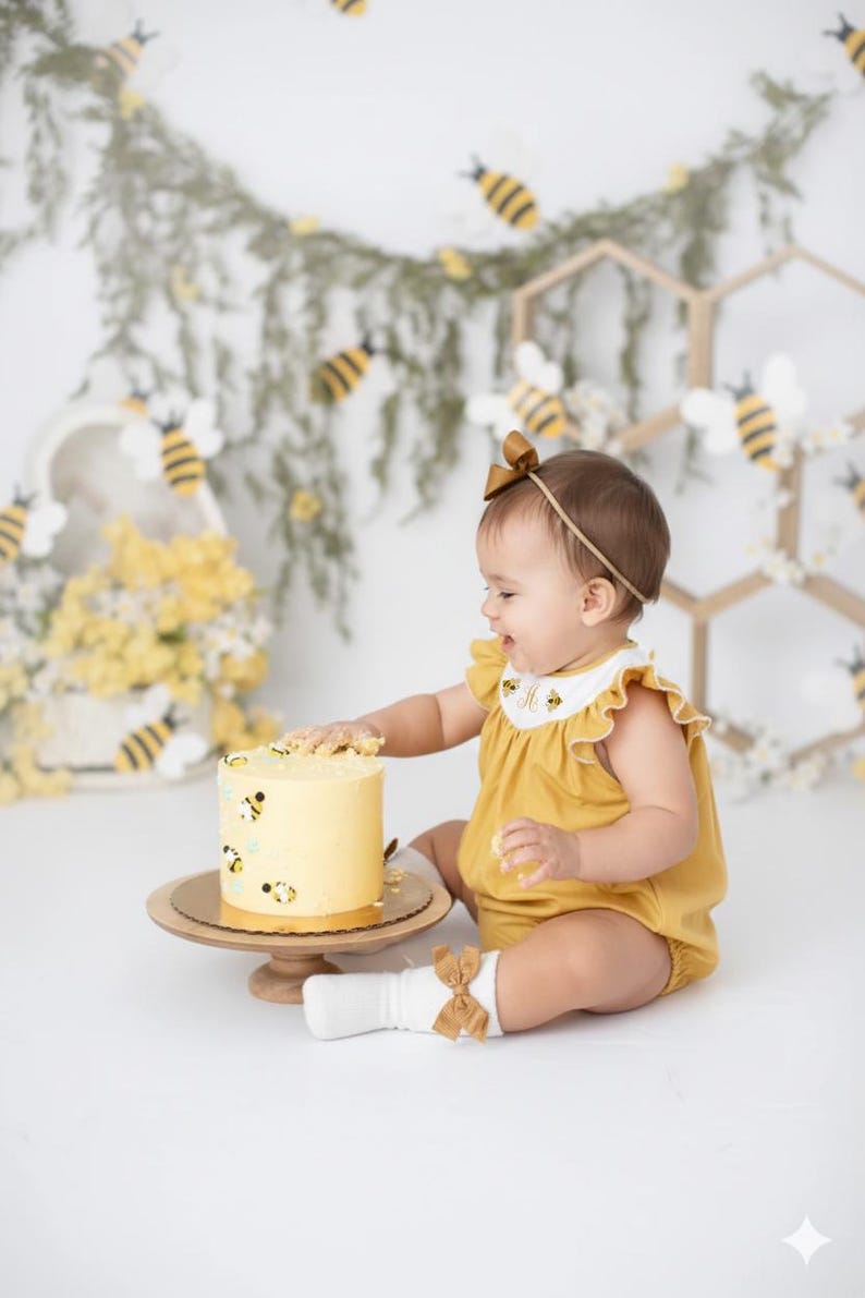 May include: A baby in a mustard yellow romper with a matching headband and socks, sits near a yellow cake decorated with bee illustrations. The cake sits on a wooden cake stand. The background includes bee decorations.