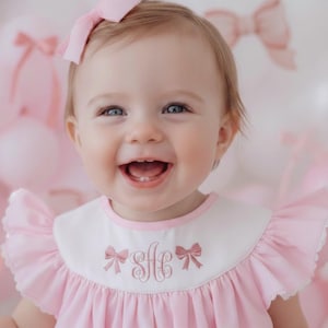 May include: A smiling baby girl wearing a pink dress with a white collar and ruffled sleeves. The collar features embroidered bows and initials. She has a pink bow in her hair, with a background of pink balloons.