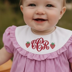 May include: A smiling baby wearing a pink gingham dress with a white collar. The collar is embroidered with red initials and strawberries. A large pink bow adorns the baby's head.