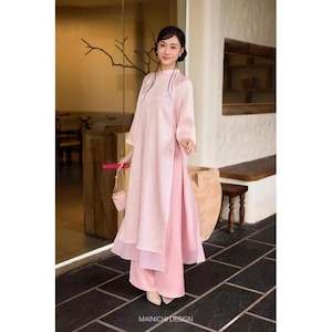 Op de afbeelding: Een lichtroze jurk met lange mouwen, een hoge kraag en een bijpassende broek. De jurk heeft een bloemenpatroon en is gelaagd over een lichtere roze rok. Een kleine roze handtas wordt vastgehouden. De tekst "MAINICHI DESIGN" staat onderaan.