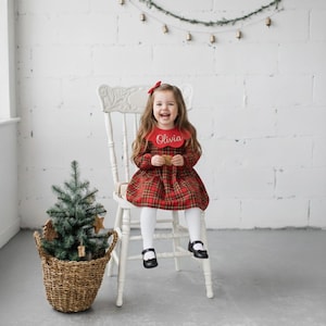 May include: A young child wearing a red plaid dress and a bib with the name "Olivia" embroidered on it. The child is sitting on a white chair. A small Christmas tree is in a woven basket.