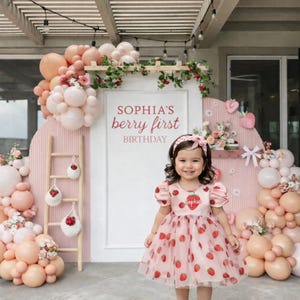Pode incluir: Uma menina com um vestido rosa com tema de morango sorri em frente a um cenário de aniversário. O cenário apresenta balões, flores e o texto "SOPHIA'S berry first BIRTHDAY". Uma faixa de cabelo a condizer completa o look.