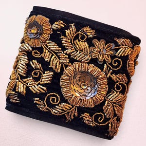May include: A black fabric cuff featuring intricate gold-toned embroidery and metallic thread work. The design includes floral motifs and leaf patterns accented with circular sequins and beads, creating a textured surface across the entire piece.