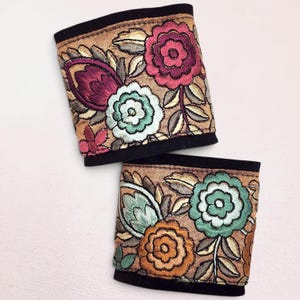 May include: Two rectangular fabric cuffs featuring intricate floral embroidery on a tan background. The designs include magenta, mint green, and orange flowers with metallic gold-toned leaves, all bordered by solid black fabric edges.