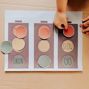 May include: A child's hand placing a red circle on a traffic light learning tool. The printed sheet features three traffic lights with circles in red, yellow, and green. The words "SLOW" and "GO" are printed on the sheet.