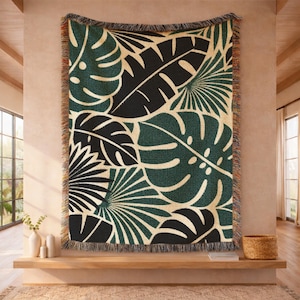 May include: A woven throw blanket featuring a tropical leaf pattern in shades of green, black, and cream. The blanket has a fringed edge and is displayed on a wall, adding a touch of nature-inspired decor to the room.