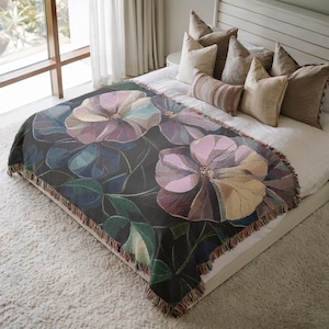 May include: A dark floral throw blanket with large, stylized flowers in shades of purple, pink, and beige. The blanket has a fringed edge and is draped over a bed. The flowers are set against a dark background with green leaves.