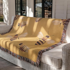 May include: Mustard yellow throw blanket featuring a repeating pattern of detailed honeybees with outstretched wings. The blanket has a fringed edge with a mix of colors. The blanket is draped over a white wicker couch.