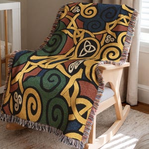 May include: A woven throw blanket featuring a repeating pattern of gold, green, dark blue, and rust-colored interlocking Celtic knots and spiral motifs. The rectangular blanket has a fringed edge along the sides and is draped over a wooden chair.
