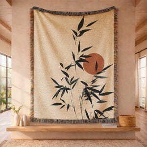 May include: Beige throw blanket with a black bamboo design and a rust-colored sun. The blanket has a fringed edge with a mix of colors. The throw is hanging on a wall above a wooden shelf.