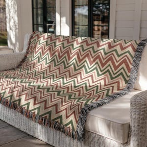 May include: A woven throw blanket with a zig-zag pattern in shades of cream, green, and burgundy. The blanket is draped over a white wicker sofa, with fringed edges. The design is a classic chevron pattern.
