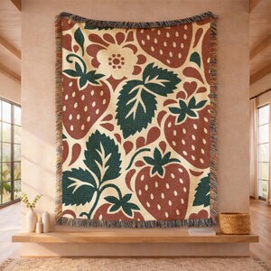 May include: A woven throw blanket featuring a repeating pattern of red strawberries, dark green leaves, and cream-colored flowers on a light beige background. The blanket has a textured knit construction and frayed edges along the top and bottom.