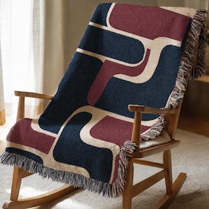 May include: A woven throw blanket featuring a geometric pattern of interlocking shapes in deep navy blue, dark burgundy, and cream. The rectangular textile has a textured surface and is finished with short, multi-colored fringe along the edges.