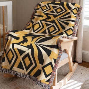 May include: A woven throw blanket featuring a bold geometric pattern in black and gold. The design consists of repeating diamonds, triangles, and angular lines. The blanket has a fringed edge with multi-colored threads along the sides.