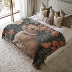 May include: A woven throw blanket featuring a detailed illustration of a brown rabbit surrounded by flowers and foliage in shades of orange, green, and cream, set against a dark background. The blanket has a fringed edge and is displayed on a bed.